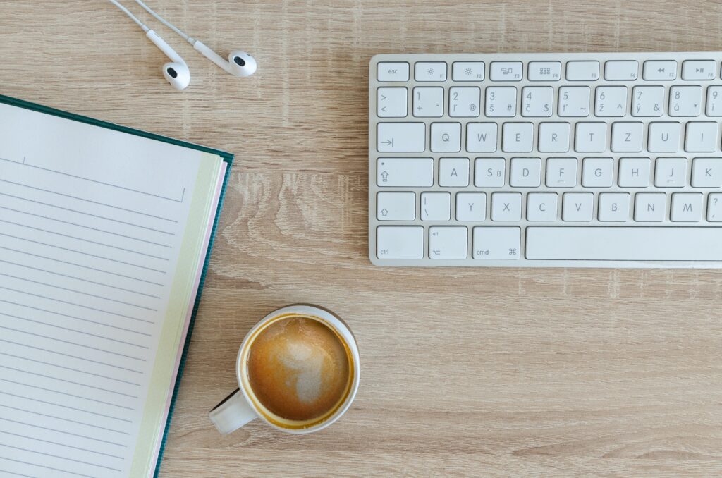 A desk with a keyboard, notebook, headphones and a cup of coffee